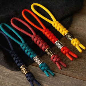 Op de afbeelding: Vijf kleurrijke paracord sleutelhangers met metalen kralen met verschillende Noorse runen. De sleutelhangers zijn in tinten geel, oranje, rood, donkerrood en teal.
