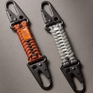 May include: Two keychains featuring black metal carabiner clips at both ends. One keychain has a brown and dark brown braided paracord strap with a small rectangular leather tag embossed with the letters ND. The other has a gray braided paracord strap.