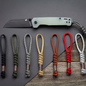 May include: A folding knife with a black blade and a pale green handle is shown above seven colourful paracord lanyards. The lanyards are in black, green, grey, tan, brown, red, and white.