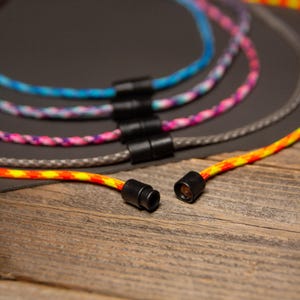 May include: Close-up of several colorful rope necklaces with black clasps. The necklaces feature vibrant patterns in shades of blue, pink, orange, and gray. The background is a dark gray surface and a wooden surface.