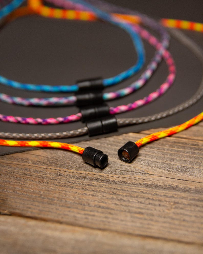 May include: Close-up of several colorful rope necklaces with black clasps. The necklaces feature vibrant patterns in shades of blue, pink, orange, and gray. The background is a dark gray surface and a wooden surface.