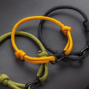 May include: Three rope bracelets in yellow, olive green, and black. Each bracelet features a knot and a black metal clasp. The bracelets are displayed on a dark gray surface.