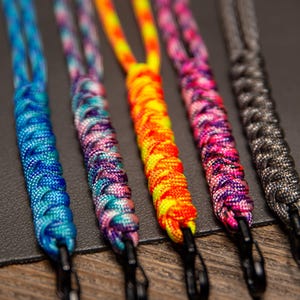 May include: Close-up of five colorful lanyards. The lanyards are made of braided cord in various colors, including blue, purple, orange, pink, and gray. Each lanyard has a black metal clip at the end.