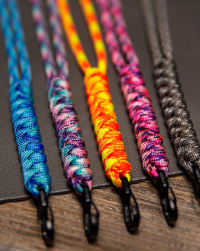 May include: Close-up of five colorful lanyards. The lanyards are made of braided cord in various colors, including blue, purple, orange, pink, and gray. Each lanyard has a black metal clip at the end.