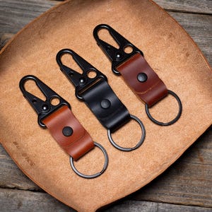 May include: Three black metal keychains with leather straps. The straps are brown and have a dark brown accent. The keychains have a spring-loaded clip and a metal ring.