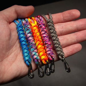 May include: A collection of six colorful paracord lanyards with black metal carabiner clips. The lanyards are in shades of blue, purple, yellow, orange, pink, and gray. Each lanyard is approximately 6 inches long.
