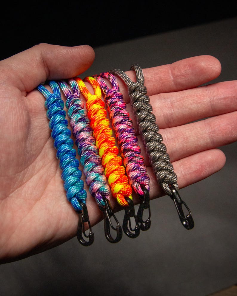 May include: A collection of six colorful paracord lanyards with black metal carabiner clips. The lanyards are in shades of blue, purple, yellow, orange, pink, and gray. Each lanyard is approximately 6 inches long.