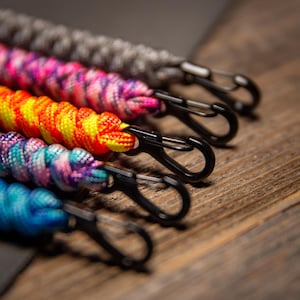 May include: Close-up of five colorful paracord lanyards with black carabiner clips. The lanyards are in shades of gray, pink, orange, purple, and blue. The background is a wooden surface.