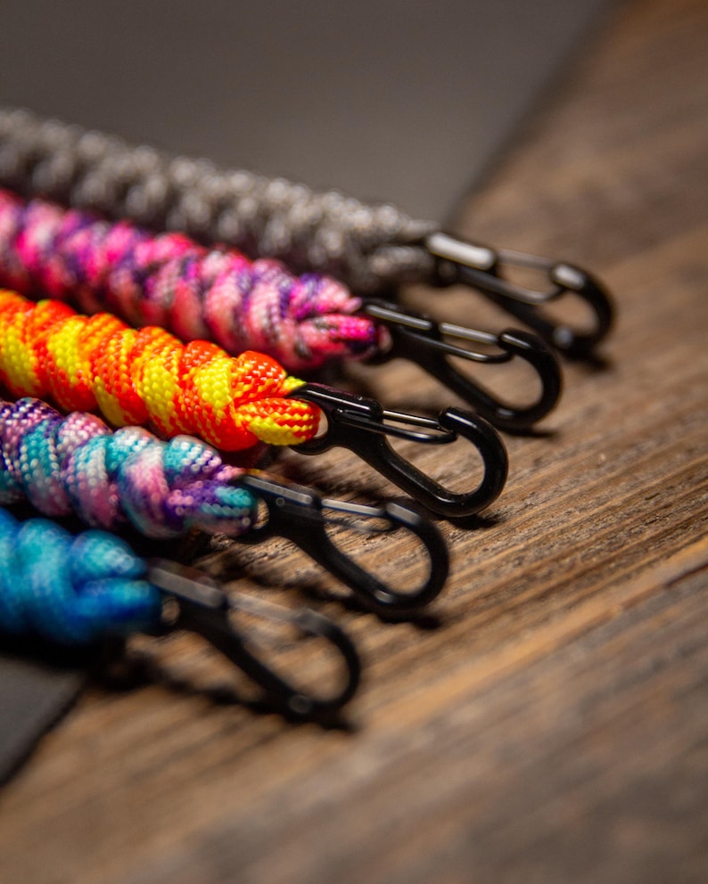 May include: Close-up of five colorful paracord lanyards with black carabiner clips. The lanyards are in shades of gray, pink, orange, purple, and blue. The background is a wooden surface.