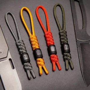 May include: Four paracord lanyards in gray, yellow, red, and olive green, each with a black bead marked "Ti". Two folding knives are visible on either side of the lanyards. The lanyards are for knives or other small items.