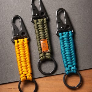 May include: Three keychains featuring braided paracord in yellow, olive green, and teal. Each keychain has a black metal carabiner clip at the top and a black metal ring at the bottom. The olive green keychain includes a small brown leather tag with the letters ND.