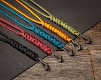 May include: A collection of colorful lanyards with metal clasps. The lanyards are in various colors, including black, red, teal, yellow, and green. The cords are tightly woven and have a textured appearance. The clasps are a silver color.