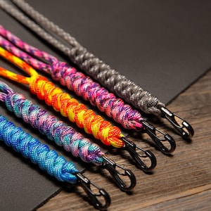 May include: Close-up of five colorful paracord lanyards with black metal clips. The lanyards are in shades of blue, purple, orange, pink, and gray. The lanyards are braided and lie on a dark surface with a wooden background.