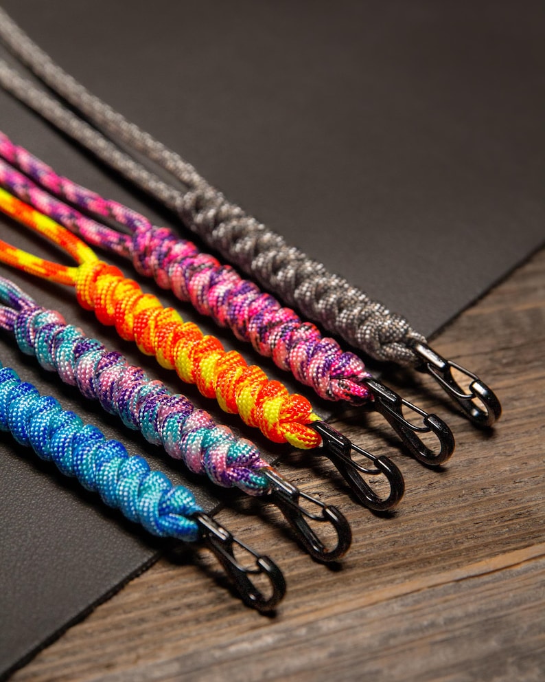May include: Close-up of five colorful paracord lanyards with black metal clips. The lanyards are in shades of blue, purple, orange, pink, and gray. The lanyards are braided and lie on a dark surface with a wooden background.