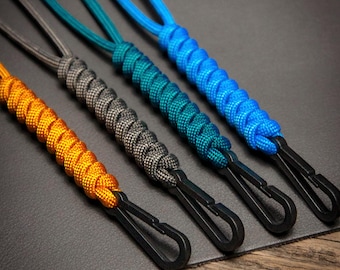 May include: Four paracord lanyards in orange, gray, teal, and blue. Each lanyard features a woven design and a black metal clip. The lanyards are displayed on a dark gray surface, showcasing their vibrant colors and textures.