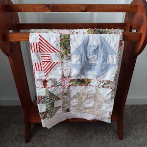 Puede incluir: Un perchero de madera para colchas con una colcha de retazos colgada. La colcha está hecha de varias telas en tonos rojos, blancos, azules y verdes. La colcha está decorada con pompones rosas.