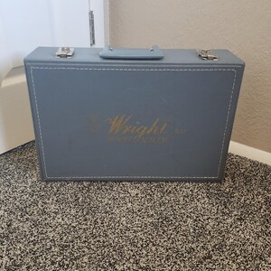 Op de afbeelding: Een blauwgrijze rechthoekige koffer met een handvat en twee zilveren sluitingen. De koffer heeft witte stiksels rond de randen en gouden letters met de tekst "Wright Way BEAUTY ACADEMY". De koffer staat op een gespikkelde vloerbedekking.