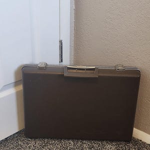 May include: A brown briefcase with silver-colored accents. The rectangular case has a handle and two latches. The briefcase is resting on a speckled carpet, near a white door and a beige wall.