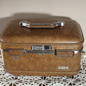 May include: A rectangular brown hardshell train case with a textured finish, featuring a silver-toned metal handle on top, a central three-digit combination lock, and two silver-toned latches. The case has small feet on the bottom base.