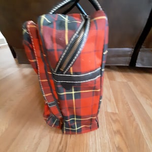 May include: A red, black, and yellow plaid travel bag with a black leather handle and zipper. The bag is standing upright on a wooden floor.