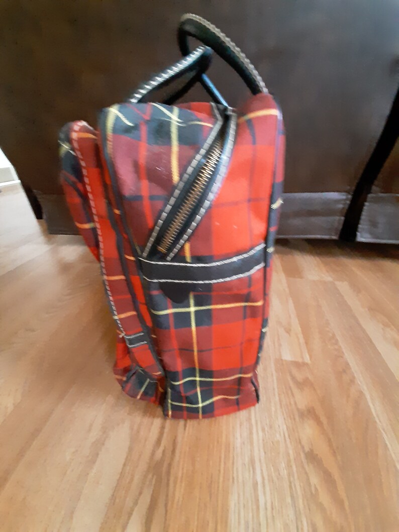 May include: A red, black, and yellow plaid travel bag with a black leather handle and zipper. The bag is standing upright on a wooden floor.
