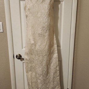 May include: A sleeveless, ivory-colored dress with a high, rounded neckline. The dress has a textured, all-over floral pattern and a straight, knee-length hemline. The dress is displayed on a mannequin.