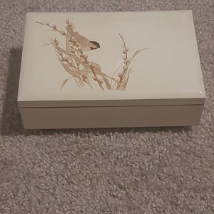 May include: A white wooden box with a delicate floral design featuring a small bird perched on a branch. The design is in shades of brown and beige.