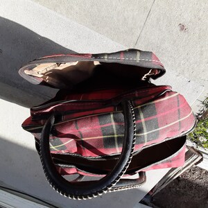 May include: A red and black plaid travel bag with a black leather handle and zipper closures.