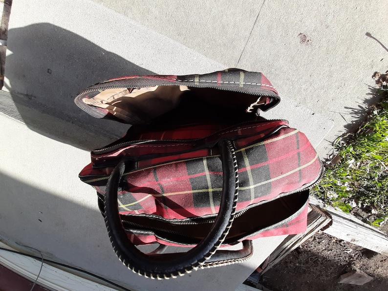 May include: A red and black plaid travel bag with a black leather handle and zipper closures.