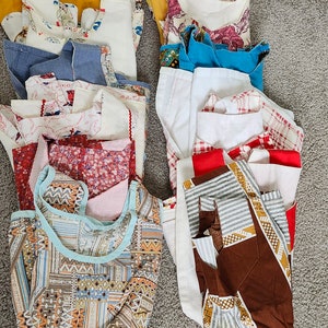 May include: A stack of vintage aprons in various colors and patterns, including floral, geometric, and striped designs. The aprons are made of different fabrics, such as cotton and linen.