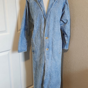 May include: Light blue denim duster coat with a classic collar and long sleeves. The coat features two front pockets and gold-colored buttons. The longline design extends to the ankles. A vintage-inspired outerwear piece.