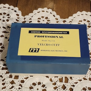 Puede incluir: Una caja azul con una etiqueta amarilla. La etiqueta dice "ANEROID SPHYGMOMANOMETER PROFESSIONAL VELCRO-CUFF Model No.115" e incluye el logotipo de Marshall Electronics, Inc. La caja está sobre un tapete de encaje blanco.