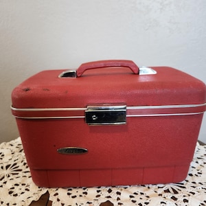 Puede incluir: Un neceser rojo vintage con ribete y asa plateados. El estuche rectangular tiene una superficie texturizada y un cierre plateado. El estuche está sobre un mantelito de encaje blanco.