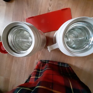 May include: Two white thermal beverage containers with red lids. The containers are empty and the lids are off. The containers are sitting on a wooden surface.