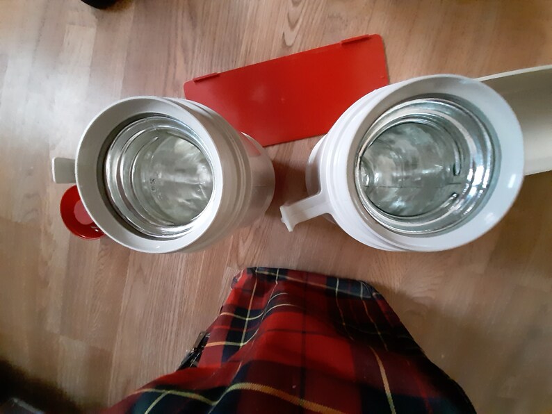 May include: Two white thermal beverage containers with red lids. The containers are empty and the lids are off. The containers are sitting on a wooden surface.