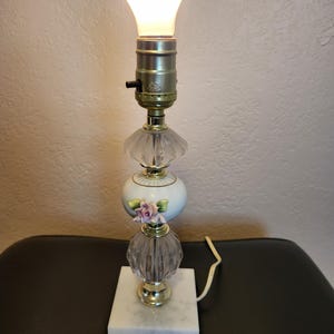 May include: A small table lamp with a white porcelain base decorated with pink roses. The lamp has a clear glass shade and a gold-colored metal base. The lamp is sitting on a white marble base.