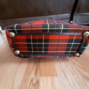 May include: A red, black, and yellow plaid bag with a black leather trim and gold metal accents. The bag is lying on a wooden surface.