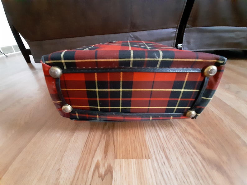 May include: A red, black, and yellow plaid bag with a black leather trim and gold metal accents. The bag is lying on a wooden surface.