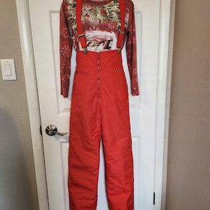 May include: A vintage ski suit comprising red salopettes with braces and a long-sleeved shirt with a winter scene print. The salopettes have a high waist and a front zip. The shirt has a red background with a winter landscape design.