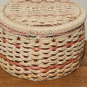Könnte beinhalten: Ein runder Nähkorb mit Deckel und gewebtem Design. Der Korb hat eine natürliche beige Farbe mit rosa Akzenten und einen Klappverschluss. Der Korb besteht aus gewebten Materialien und steht auf einer Holzoberfläche.
