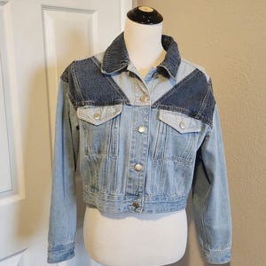May include: A light blue denim jacket with dark blue denim panels on the shoulders and sleeves. The jacket has a button closure and two front pockets.