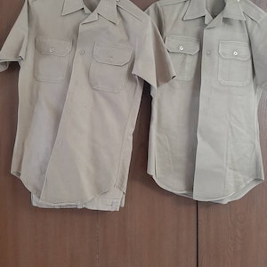 Puede incluir: Dos camisas de manga corta color caqui con cuello abotonado y bolsillos colgadas de perchas sobre un fondo de madera.