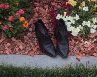 Peut inclure: Une paire de bottines en cuir noir à bout pointu, posée sur des copeaux de bois près de fleurs colorées. Les bottines ont un petit talon et une découpe en V sur le dessus.