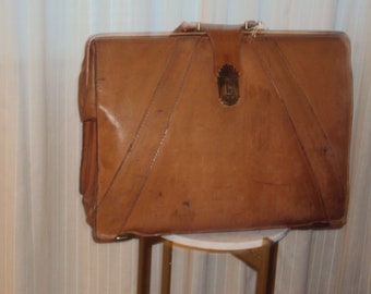 May include: A vintage brown leather suitcase with a brass latch and a strap. The suitcase is sitting on a small white table.