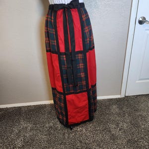 May include: A long, patchwork skirt featuring a combination of red and plaid fabric. The skirt has a black waistband with a bow and vertical black panels. The plaid incorporates green, red, and yellow hues.