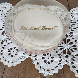 Peut inclure: Un récipient en plastique transparent vintage avec les mots "BEE HIVE" et "Pin-Curl Bonnet" en lettres dorées. Il contient un bonnet à boucles blanc. Le récipient est posé sur un napperon en dentelle blanche.