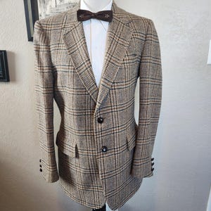Op de afbeelding: Een bruin en beige geruit jasje met een wit overhemd en een bruine vlinderdas. Het jasje heeft twee voorzakken en drie knopen op elke manchet. De outfit wordt op een mannequin gepresenteerd.