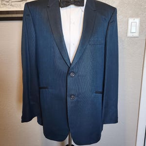 Puede incluir: Una chaqueta de esmoquin azul marino con solapa de satén negro y pajarita a juego. La chaqueta tiene cierre de dos botones y una camisa blanca debajo. El esmoquin se exhibe en un maniquí.