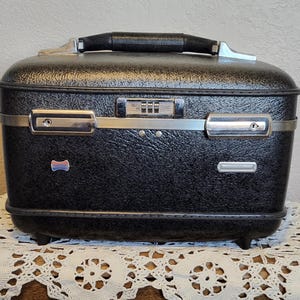 Puede incluir: Una maleta de viaje negra vintage con un exterior texturizado, detalles metálicos plateados y una cerradura de combinación. La maleta tiene un asa negra y está colocada sobre un mantel de encaje blanco.