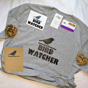 May include: A grey t-shirt with the text "Bird Watcher" and a silhouette of a bird. The shirt is packaged with a card, two stickers, and two small bags of birdseed.